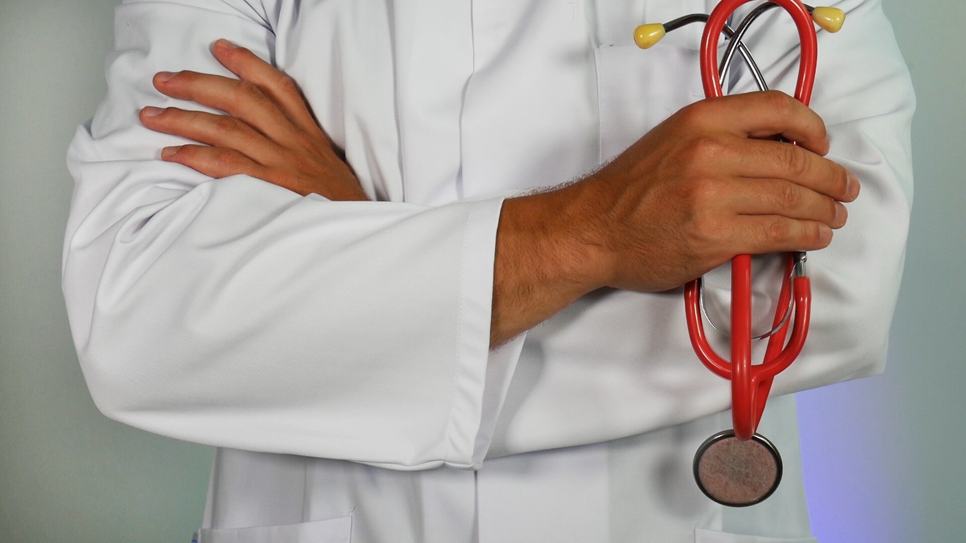 A doctor in a white lab coat with crossed arms holding a red stethoscope.