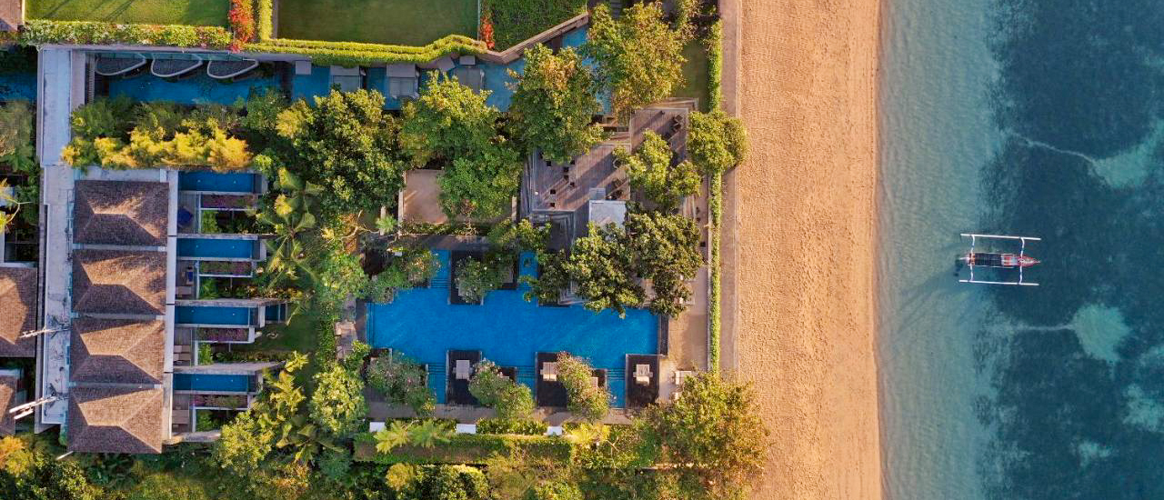 Aerial view of a beautiful resort with pools, sandy beach, and a boat in the clear ocean, Bali.