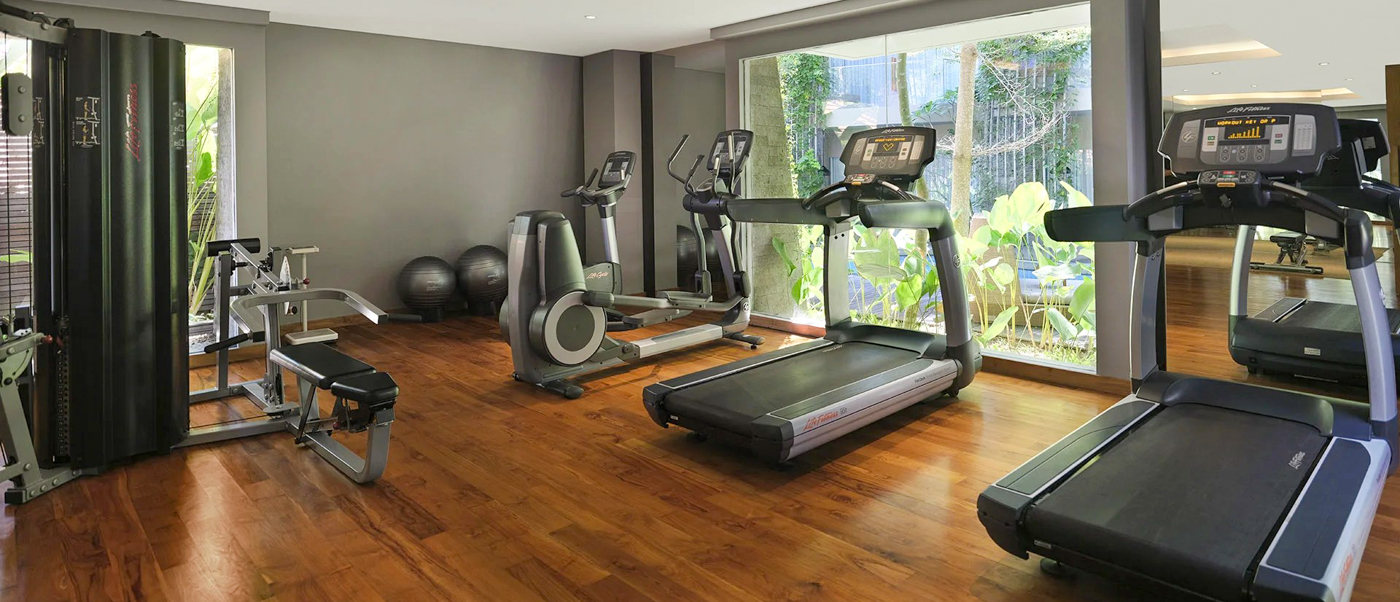 Modern hotel gym with treadmills, elliptical, and weight machines, overlooking tropical greenery in Sanur.