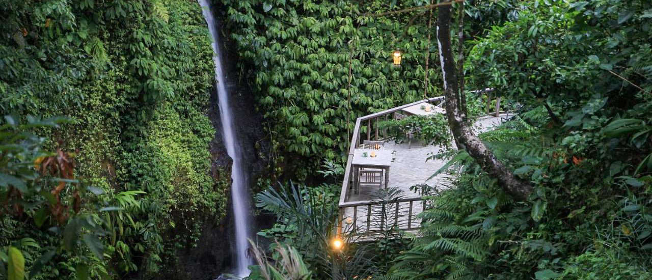 A serene waterfall cascades beside a luxurious wooden resort balcony surrounded by lush green jungle in Ubud.