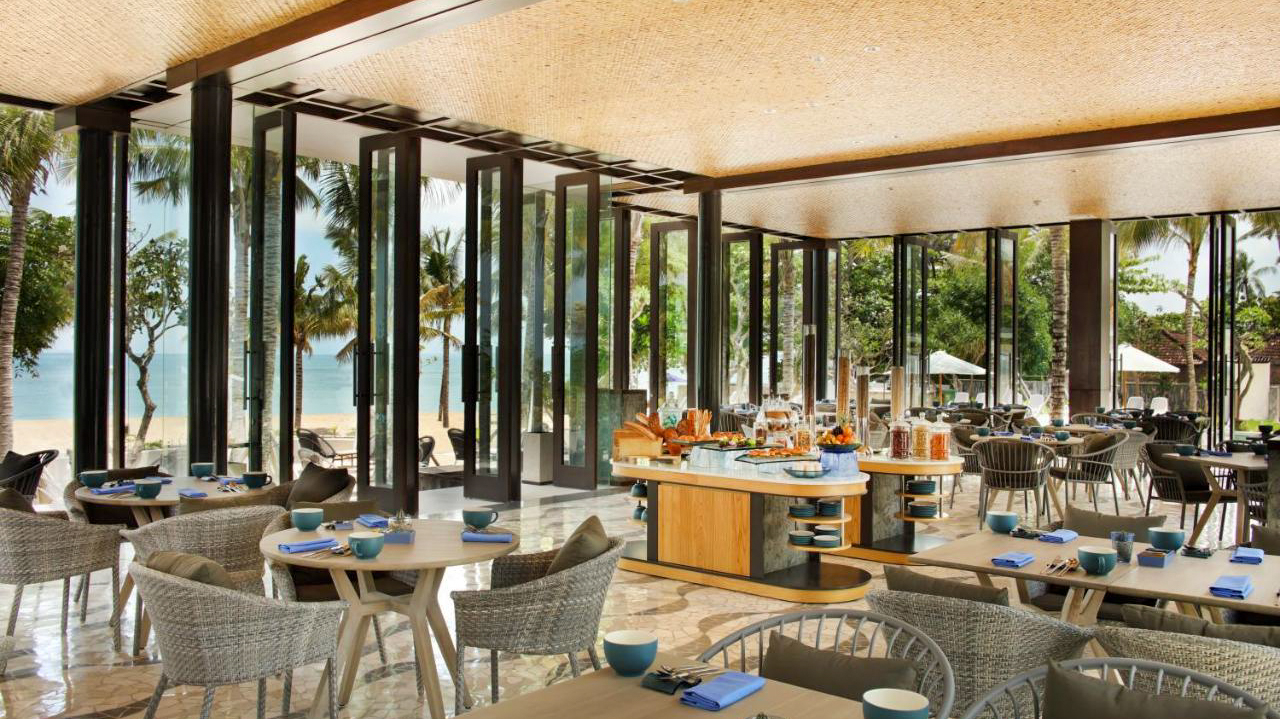 A bright, open-plan restaurant with a buffet and tables overlooking a tropical beach in Kuta.