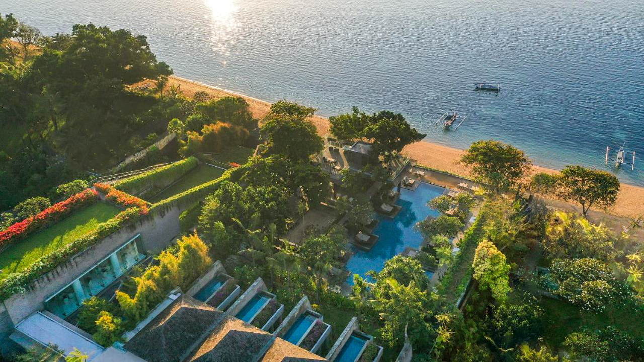 Aerial view of a luxurious tropical resort with pools, lush gardens, and a sandy beach in Bali.