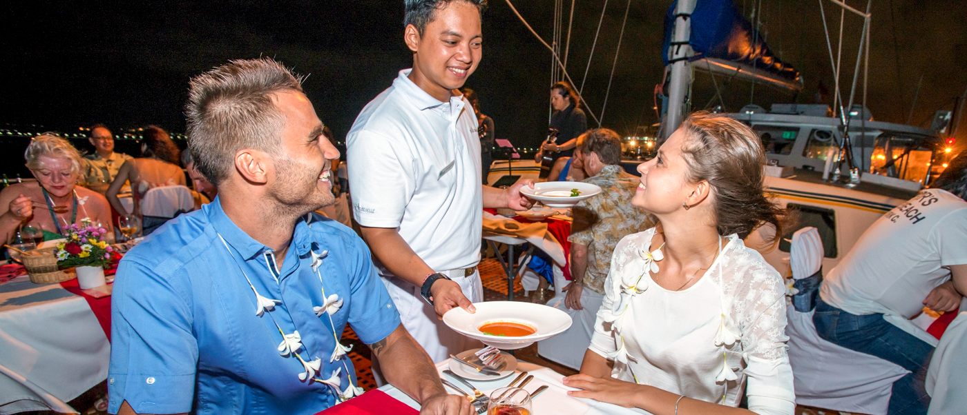 A smiling couple enjoys a romantic dinner served by staff on a luxurious evening cruise in Bali.