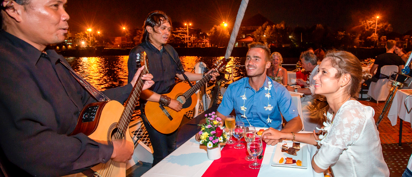 A happy couple enjoys live music and dinner on a romantic evening cruise in Benoa Harbour.