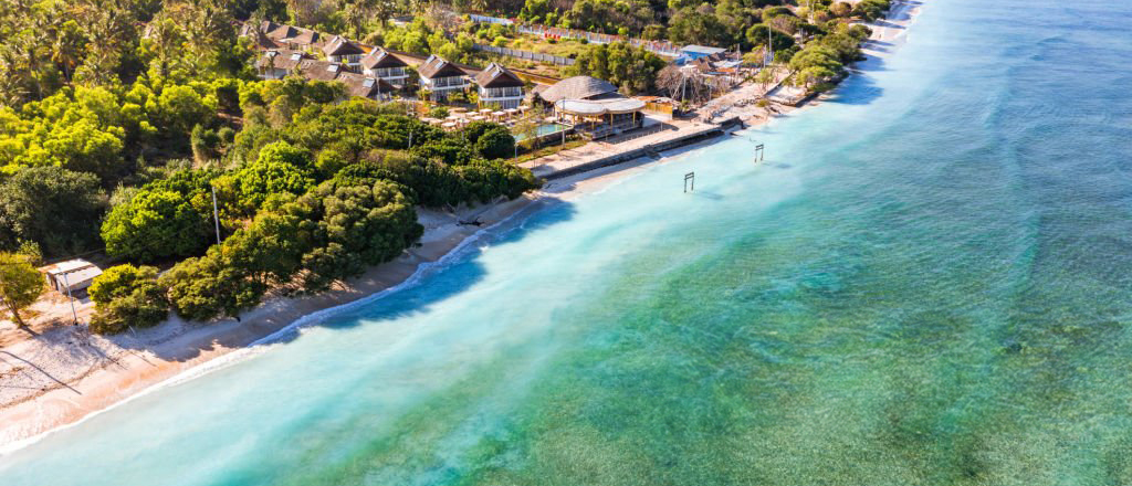Gili Trawangan's pristine sandy beach with clear turquoise waters, lush tropical trees, and beachfront resorts.