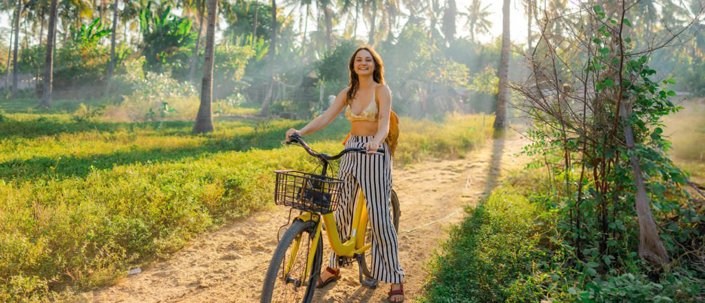 Happy woman with a yellow bike on a tropical dirt path in Gili Trawangan at sunset.