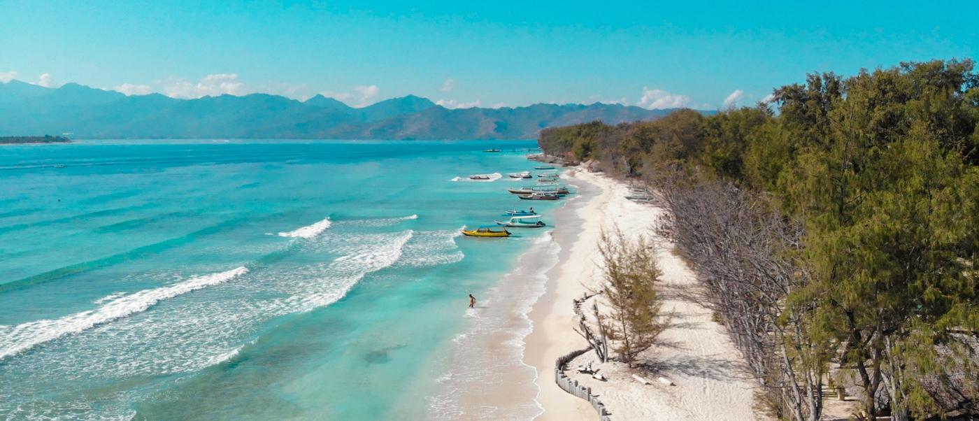 Aerial view of Gili Trawangan's pristine white sand beach, turquoise ocean, and distant mountains under a clear sky.