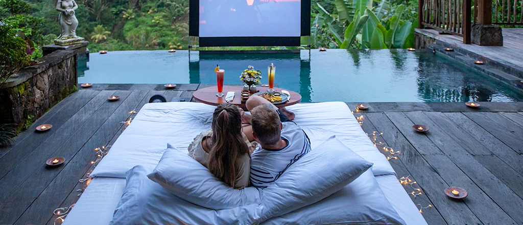 Honeymooners enjoy a romantic outdoor movie night with drinks and snacks by a pool in Bali.