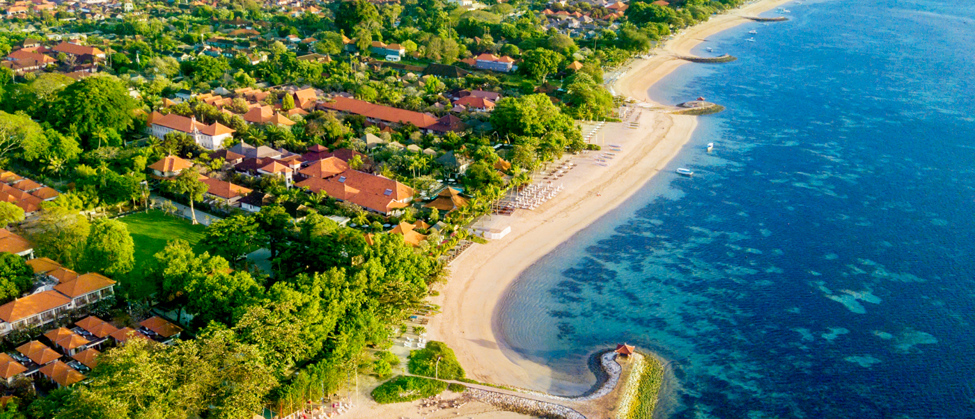 Aerial view of Sanur Beach in Bali, featuring a sandy coastline, clear blue water, and lush resorts.