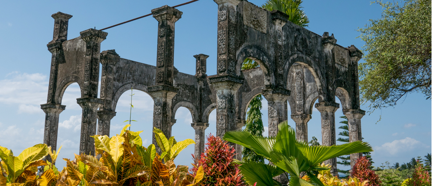 Weathered stone arches and columns of Taman Ujung Water Palace in Bali, surrounded by vibrant tropical plants.
