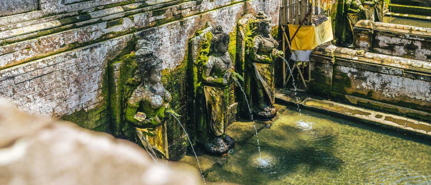 Moss-covered stone statues spout holy water into a sacred pool at Tirta Empul Temple, Bali.