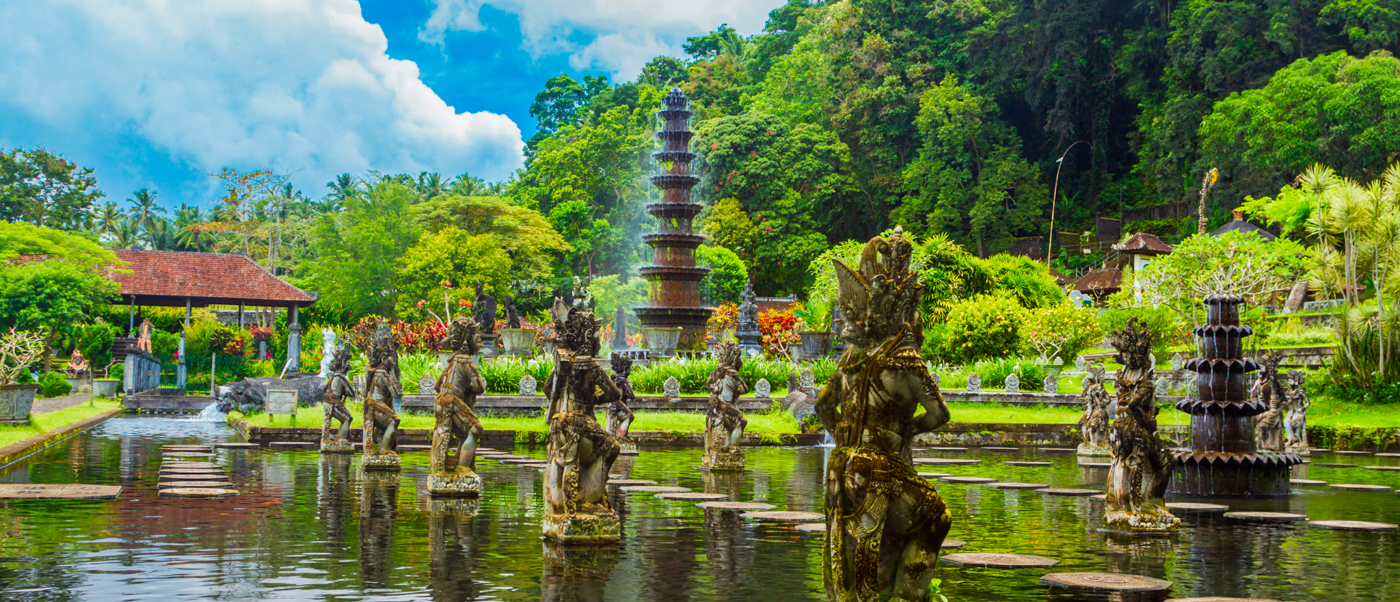 Ornate stone statues stand in the serene water garden of Tirta Gangga, Bali, under a blue sky.