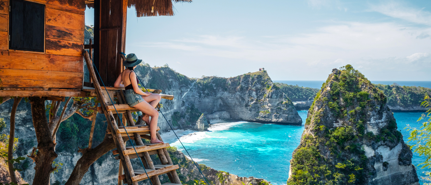Woman on a treehouse balcony overlooking the stunning turquoise ocean and dramatic cliffs of Nusa Penida.