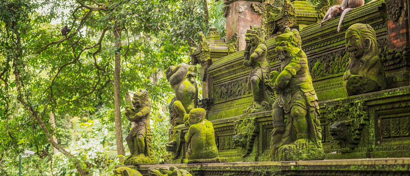 Playful monkeys perched on ancient, moss-covered stone statues amidst the lush greenery of Ubud, Bali.