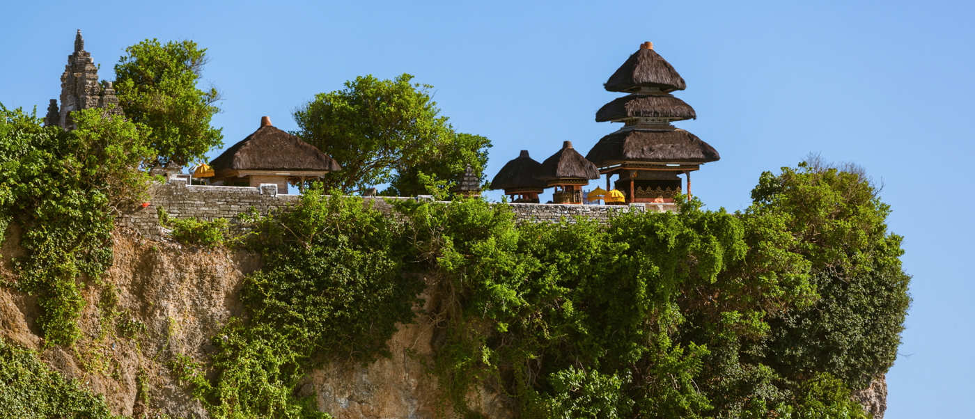 Ancient Uluwatu Temple with multi-tiered thatched roofs perched on a verdant cliff under a clear blue sky.