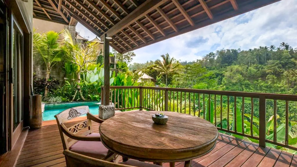 A luxurious private balcony with a wooden table and chairs, overlooking a lush Bali jungle and pool.