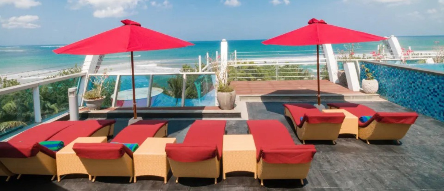 Red lounge chairs and umbrellas on a sunny rooftop terrace overlooking the ocean and Kuta Beach.