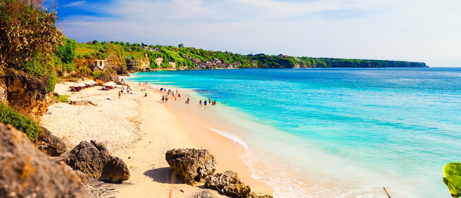 Tourists relax on the sunny white sand beach with clear turquoise water at Kuta, Bali.