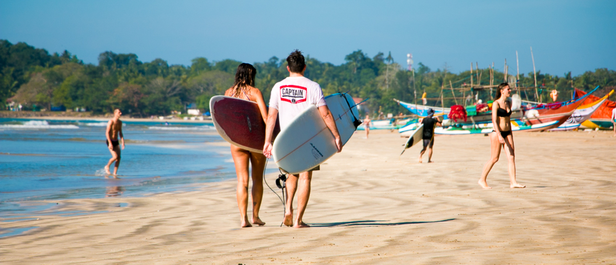 A couple walks on the golden sand of Weligama beach, carrying surfboards under a sunny sky.