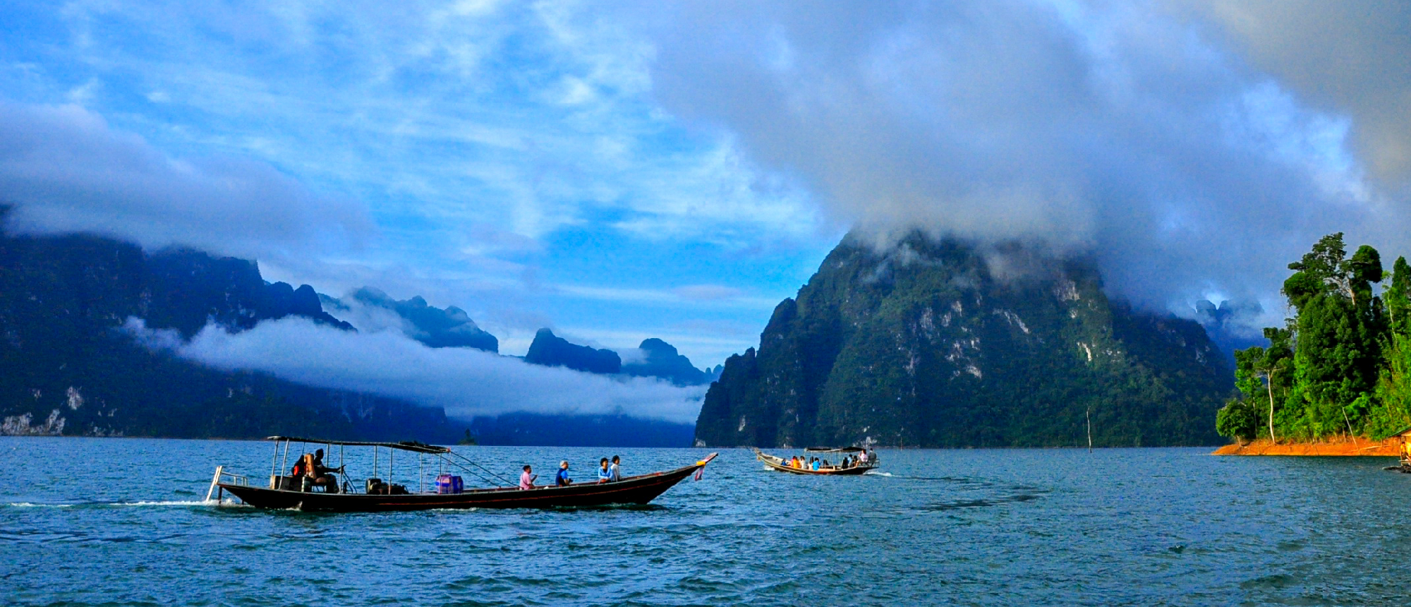 Tourists enjoy a serene boat ride past majestic, mist-shrouded limestone karsts on calm blue water.