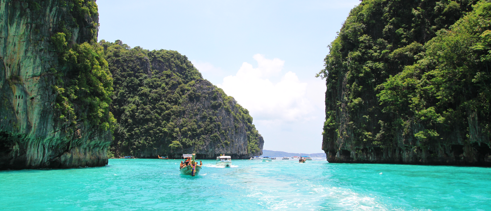 A longtail boat glides through turquoise water between towering green limestone cliffs in Phi Phi Islands.