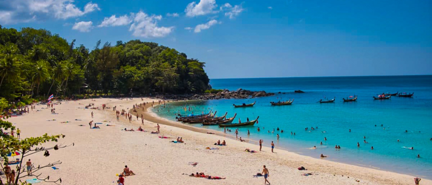 A vibrant Phuket beach with many people swimming, sunbathing, and longtail boats in turquoise water.