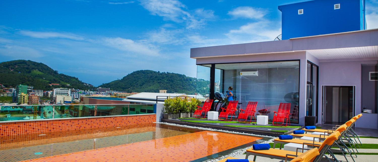 A vibrant rooftop pool with orange lounge chairs offers stunning views of Phuket's lush hills.