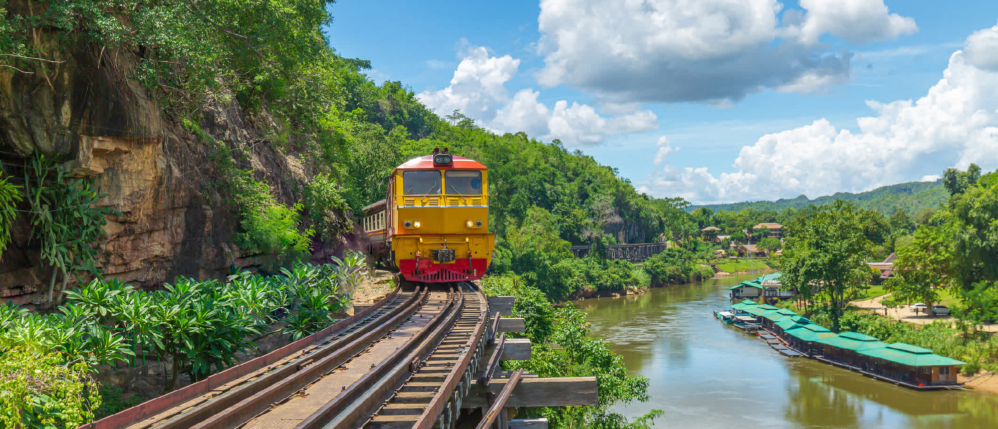A vibrant yellow and red train travels along tracks beside a scenic river, surrounded by lush Thai landscapes.