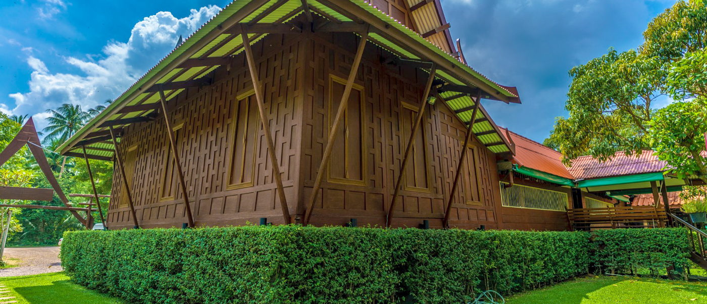 Traditional wooden Thai house with a distinctive green roof, surrounded by lush hedges in Amphawa.