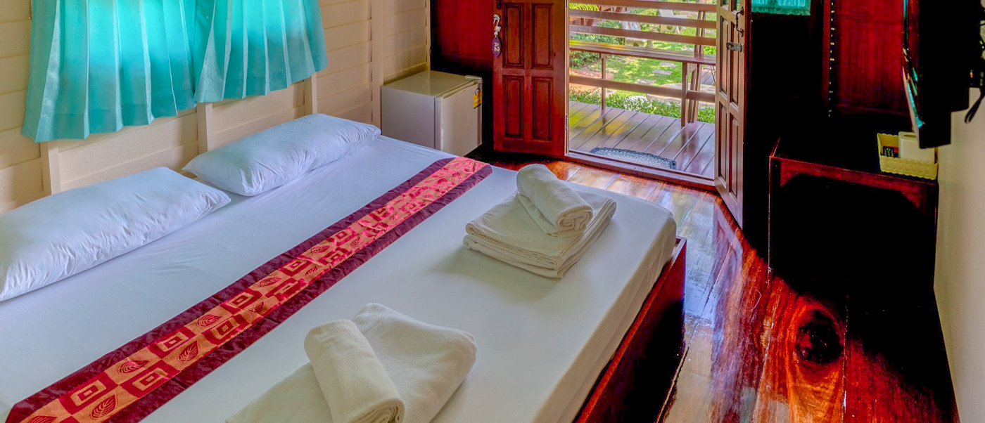 A welcoming homestay bedroom in Amphawa with a patterned bed runner and open door to greenery.