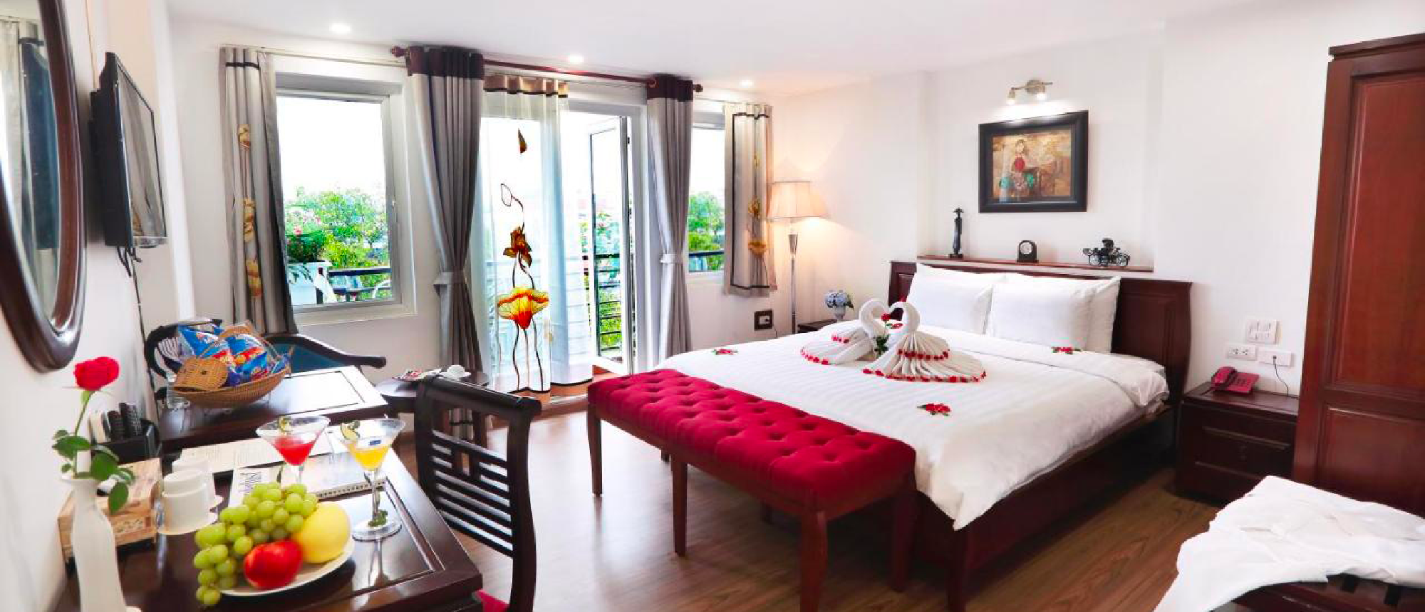 Bright hotel room in Hanoi featuring a bed with towel swans, fruit, drinks, and a balcony.