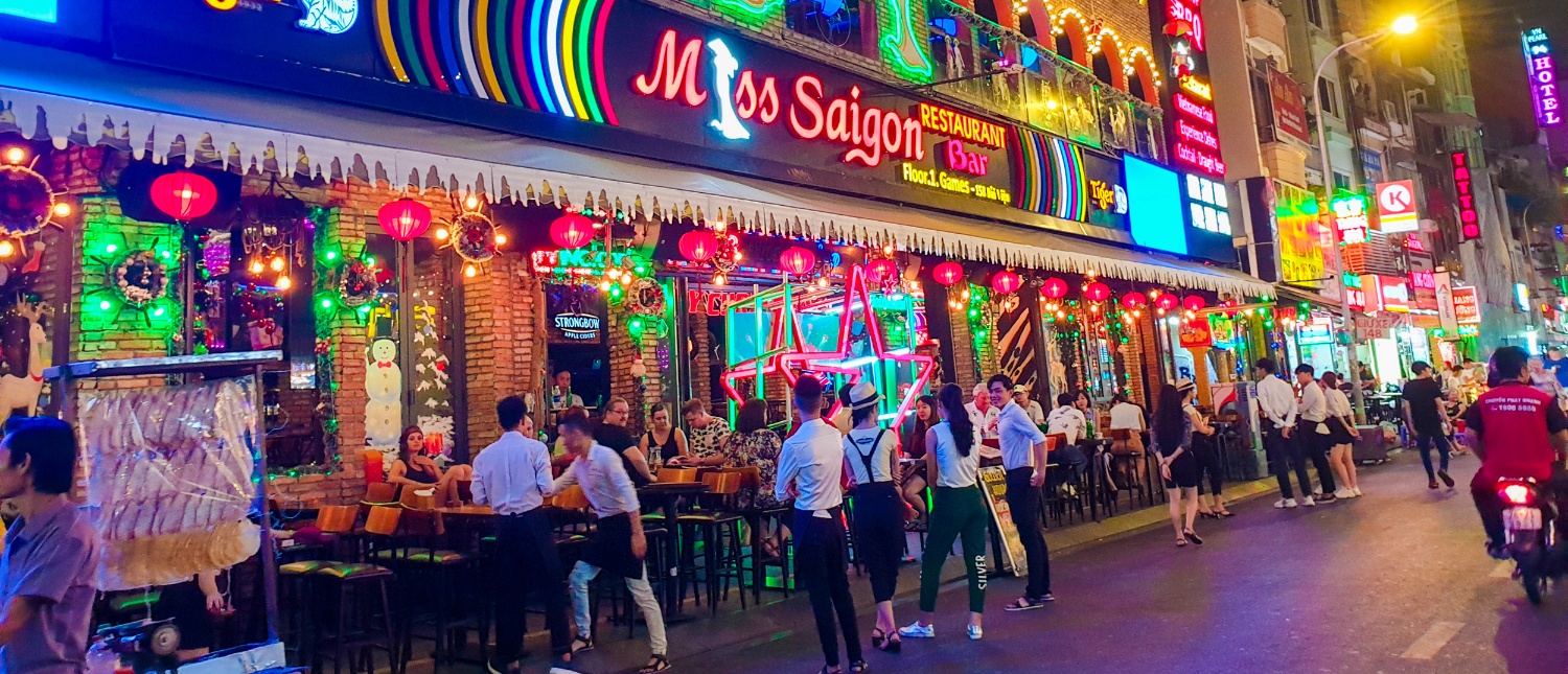 Bustling night street in Ho Chi Minh City with Miss Saigon restaurant, neon lights, and many people.