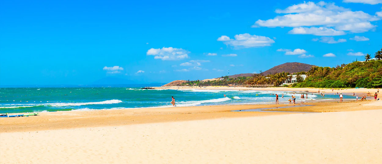 Tourists enjoy the sunny sandy beach and turquoise waves of Mui Ne, backed by green hills.
