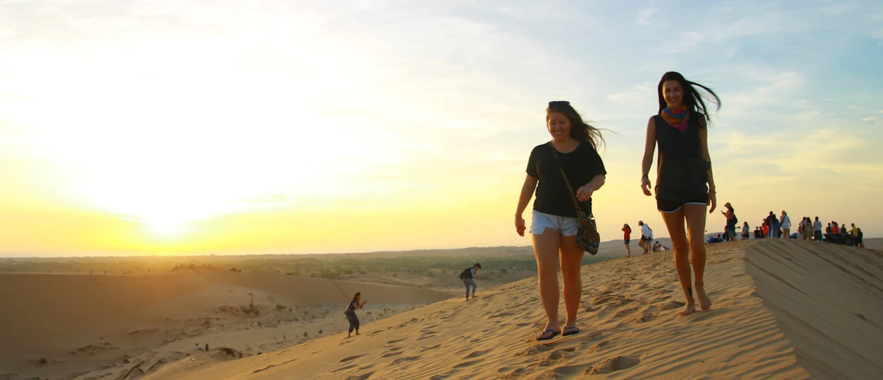 Two women descend a golden sand dune at sunrise with other visitors enjoying the desert landscape in Vietnam.
