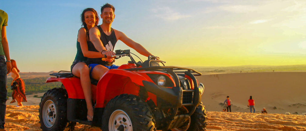 A happy couple rides a red ATV on golden sand dunes at sunrise in Vietnam.