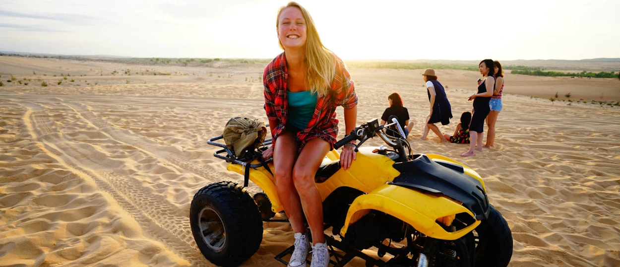 Smiling woman on a yellow ATV in the vast golden sand dunes of Vietnam at sunrise.