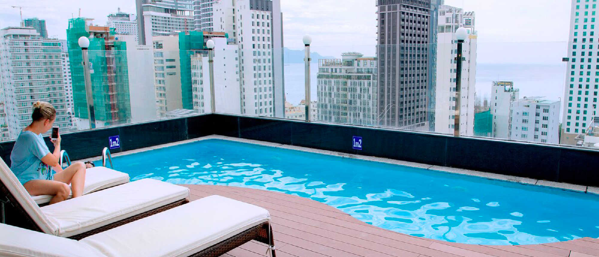 A woman relaxes by a rooftop pool, enjoying panoramic views of Nha Trang city and the ocean.