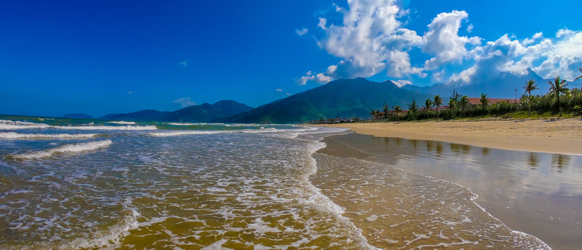 Gentle waves on the golden sand beach of Nha Trang, with mountains under a blue sky.