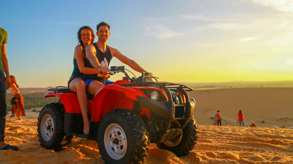 A happy couple rides a red ATV on vast sand dunes at sunset in Vietnam.