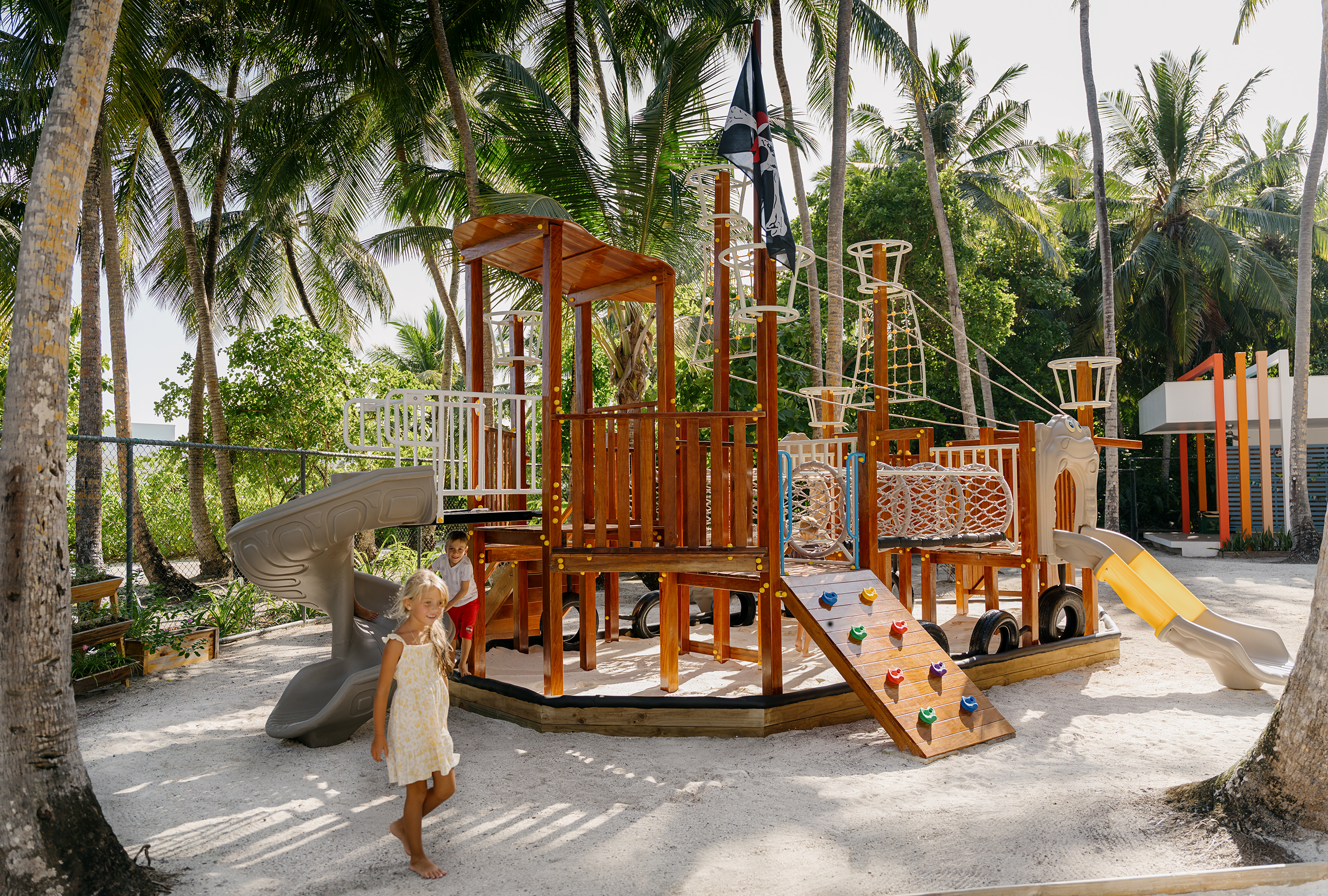Children enjoy a large pirate ship playground with slides and nets on a sandy tropical resort.