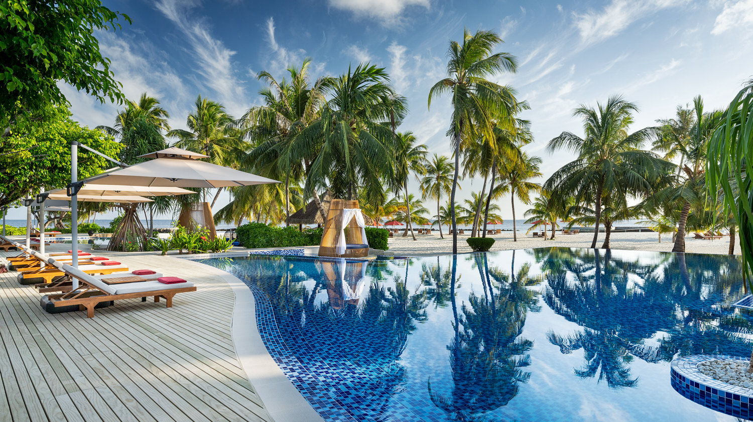 Serene resort swimming pool with lounge chairs and palm trees leading to a sandy Maldives beach.