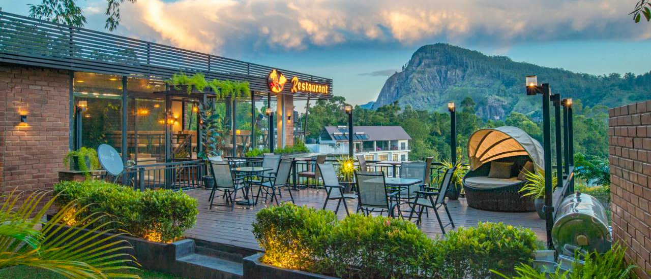 A scenic rooftop terrace with outdoor seating and lush plants, overlooking misty mountains in Ella.