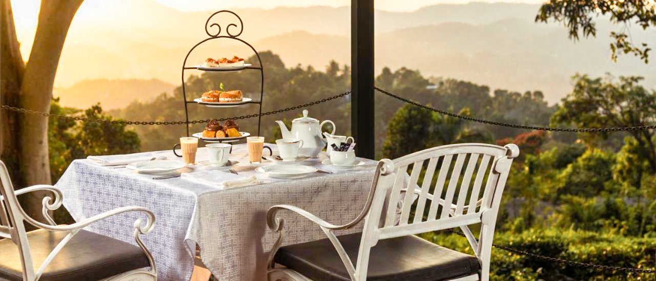 Elegant outdoor tea setting with pastries and a teapot, overlooking Kandy's lush hills at golden hour.