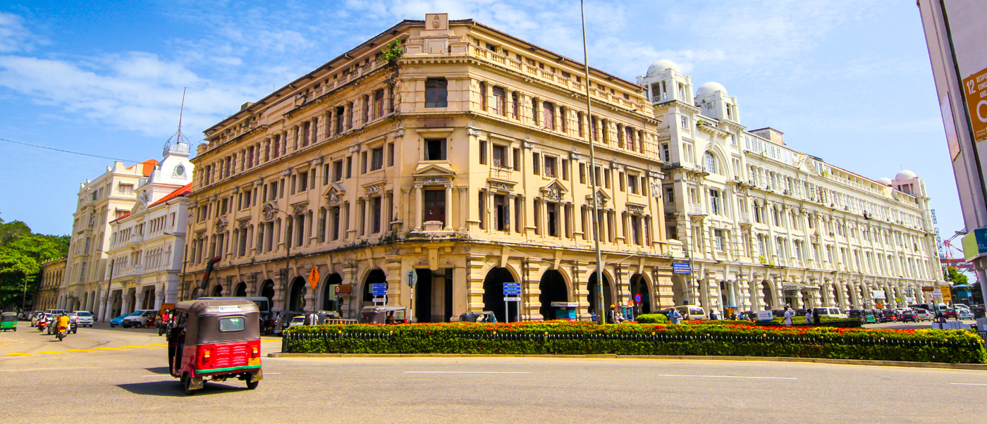 A red tuk-tuk drives past ornate colonial buildings on a sunny street in Colombo.