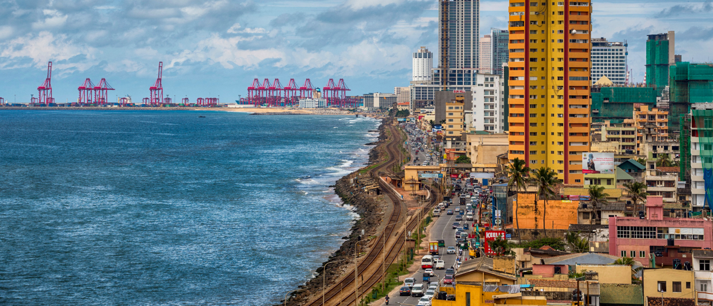 Busy Colombo coastline with a road full of traffic, colorful buildings, and a port with red cranes.