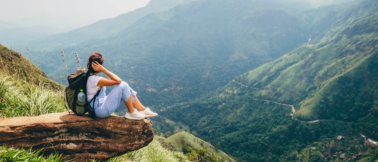 Adventurous woman with a backpack sitting on a cliff edge, enjoying sweeping valley views in Ella.