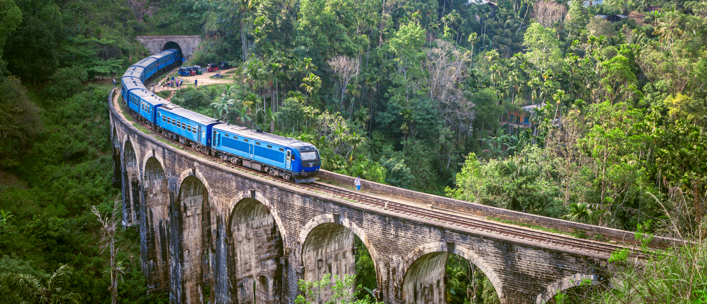 A blue train crosses the historic Nine Arches Bridge surrounded by lush green jungle in Ella, Sri Lanka.