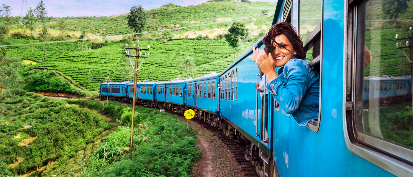 A smiling traveler leans from a blue train window, winding through lush green Sri Lankan tea fields.