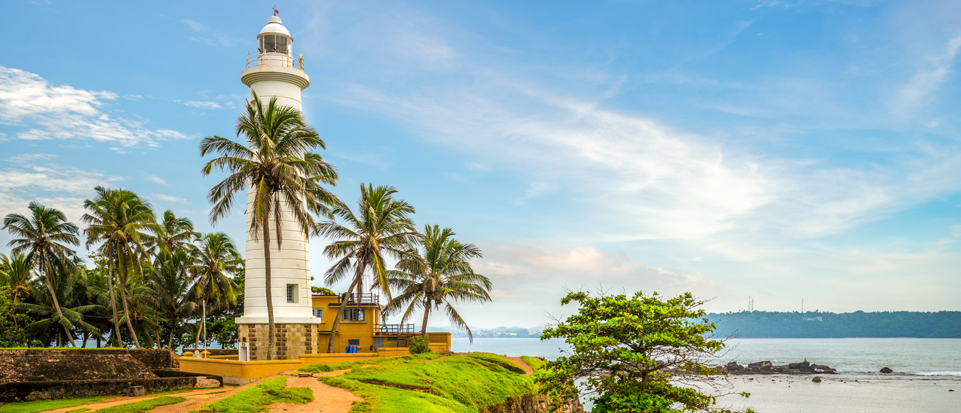 A tall white lighthouse stands among palm trees on a green cliff overlooking the sea at Galle Fort.