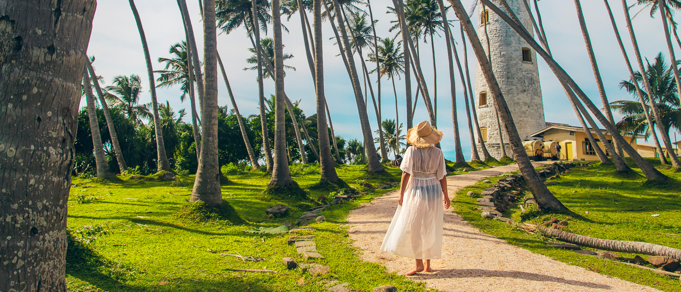 A woman in a white dress walks a palm-lined path towards a lighthouse at Galle Fort.